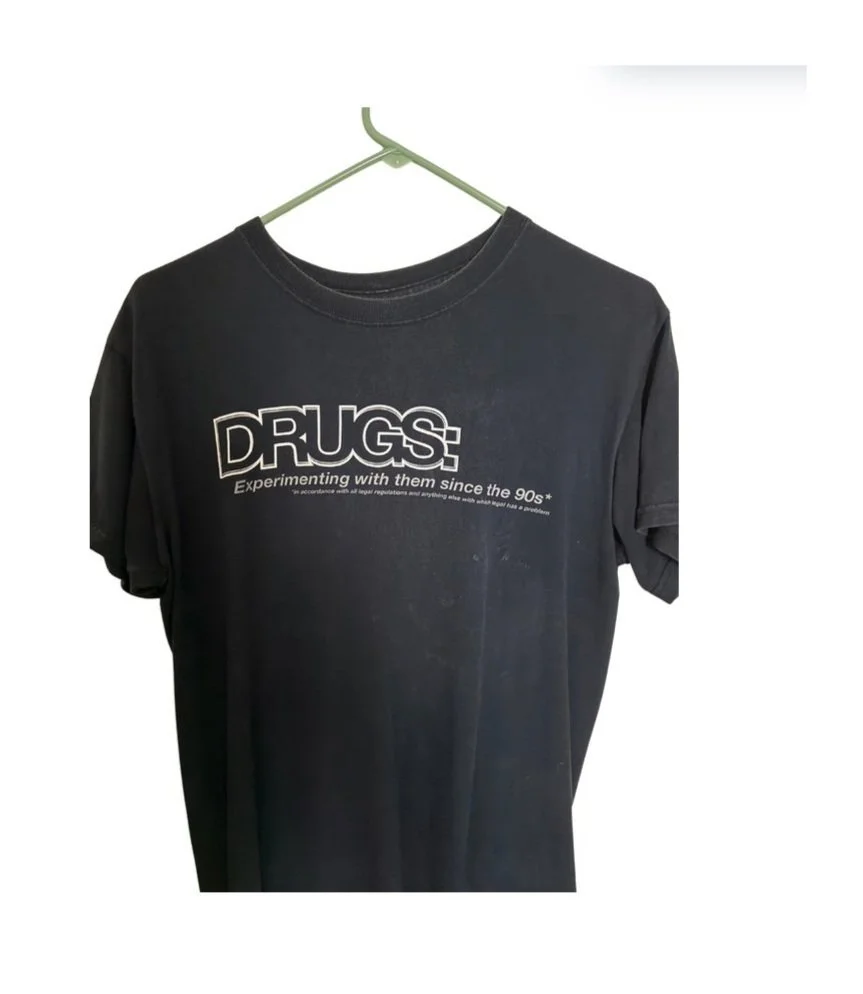 Black T-shirt hanging on a hanger with the word "DRUGS" and the phrase "Experiencing with them since the 90s" printed on it.